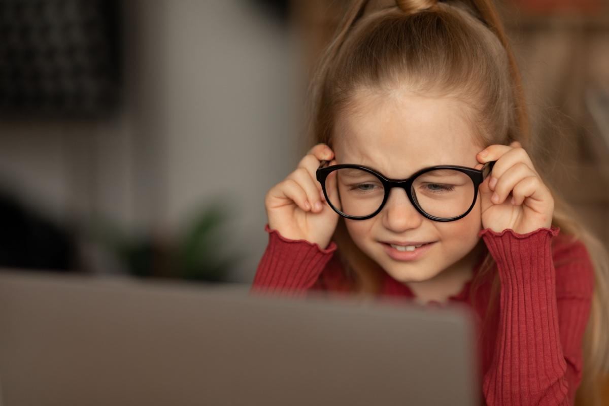 Little girl holding her glasses as she struggles to focus on her computer.